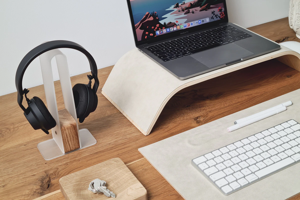 A laptop raised on a wooden stand beside headphones and an external keyboard.