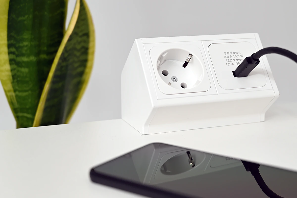 A smartphone beside a desktop power outlet and a potted plant on a white desk.